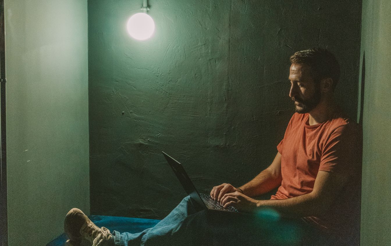 Eduardo sitting on the floor of a dark room, working on a laptop, lit by a single hanging bulb.